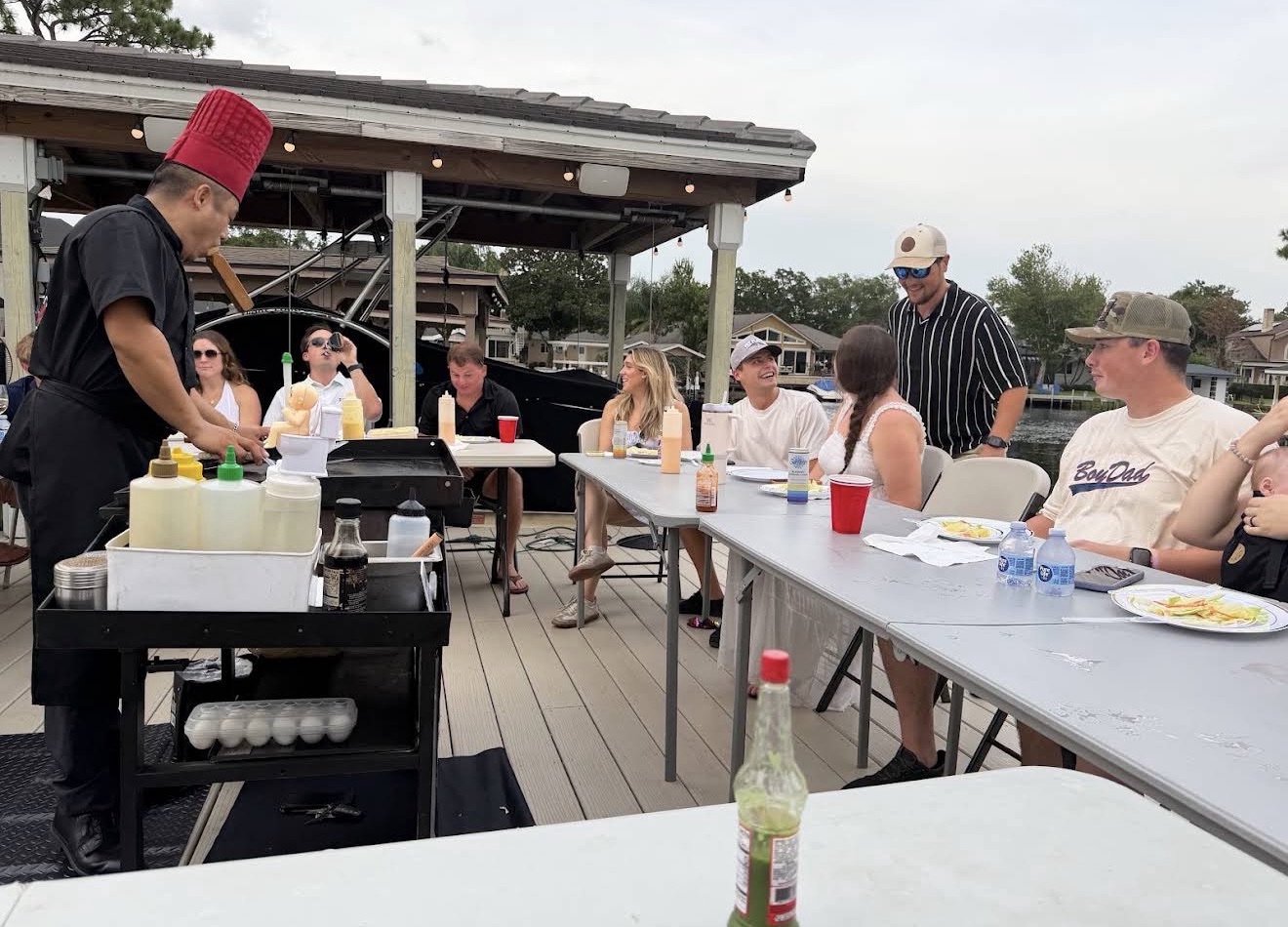 Chef in a red hat serving guests at long tables on a lakeside dock in Florida