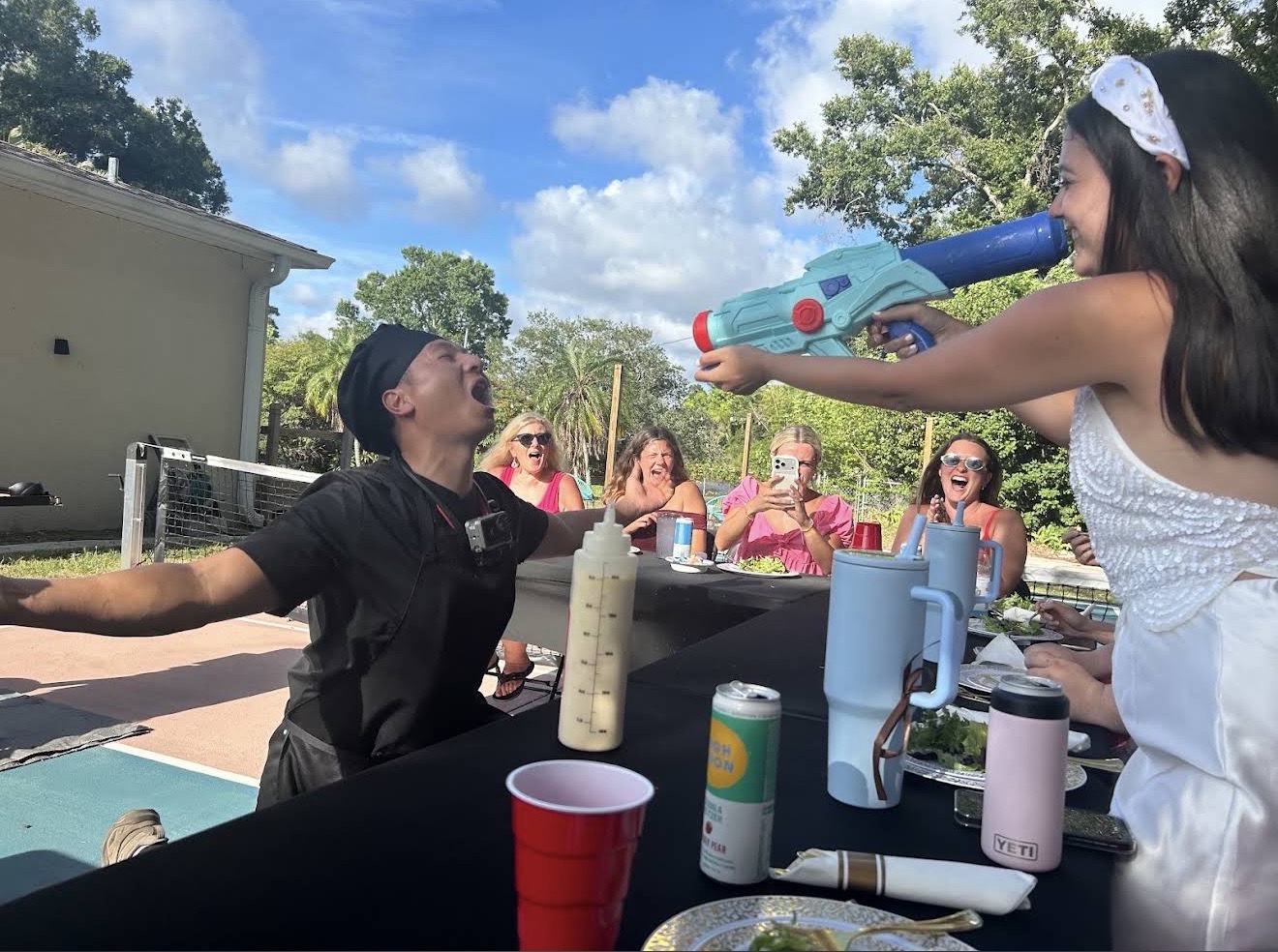 Bride playfully aiming the sake squirt gun at the laughing hibachi chef while guests watch